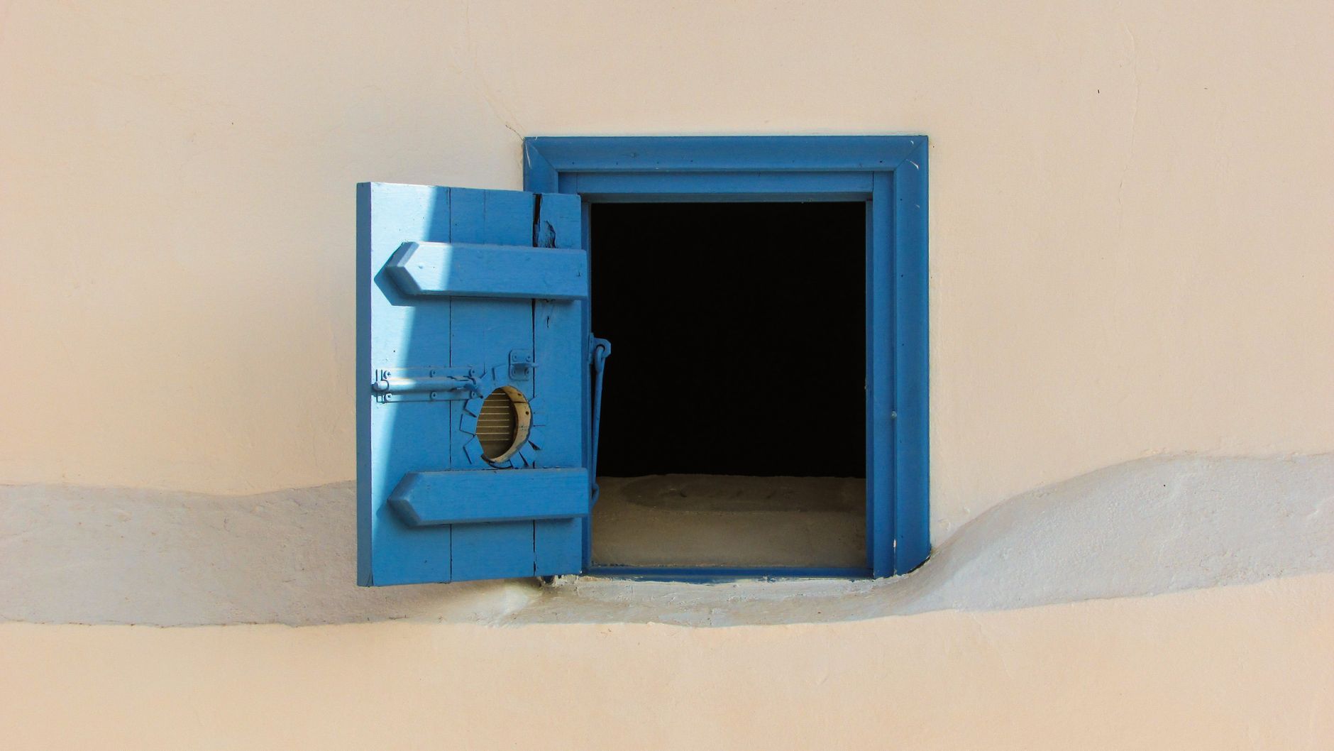 Blue wooden shutter window on a beige wall, open to reveal darkness inside